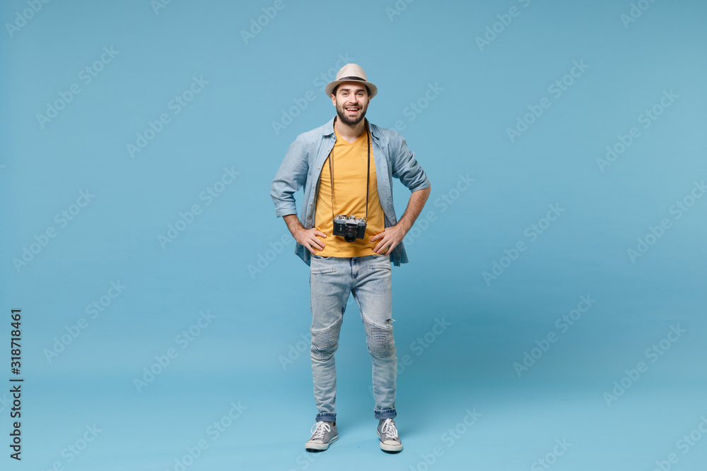 Naklejka premium Smiling traveler tourist man in yellow summer clothes with photo camera isolated on blue background. Male passenger traveling abroad on weekends. Air flight journey. Stand with arms akimbo on waist.