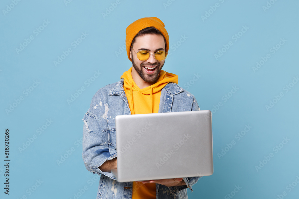 Funny young hipster guy in fashion jeans denim clothes posing isolated on pastel blue background studio portrait. People emotions lifestyle concept. Mock up copy space. Working on laptop pc computer. Funny young hipster guy in fashion jeans denim clothes posing isolated on pastel blue background studio portrait. People emotions lifestyle concept. Mock up copy space. Working on laptop pc computer.