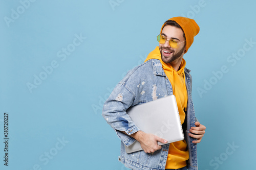 Side view of young hipster guy in fashion jeans denim clothes posing isolated on pastel blue background in studio. People lifestyle concept. Mock up copy space. Hold laptop pc computer, looking back.