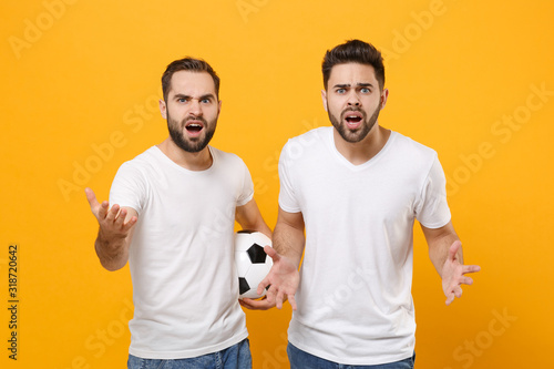 Perplexed men guys friends in white t-shirt posing isolated on yellow background. Sport leisure lifestyle concept. Mock up copy space. Cheer up support favorite team with soccer ball spreading hands. © ViDi Studio