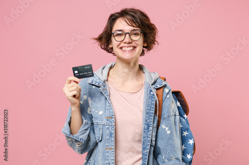 Smiling young woman student in denim clothes glasses backpack posing isolated on pastel pink background. Education in high school university college concept. Mock up copy space. Hold credit bank card.