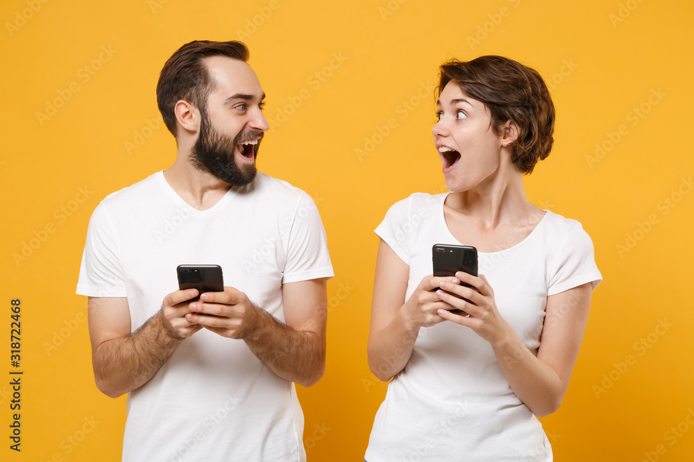 Shocked young couple friends bearded guy girl in white blank empty t-shirts isolated on yellow orange background. People lifestyle concept. Mock up copy space. Using mobile phone, typing sms message. Shocked young couple friends bearded guy girl in white blank empty t-shirts isolated on yellow orange background. People lifestyle concept. Mock up copy space. Using mobile phone, typing sms message.
