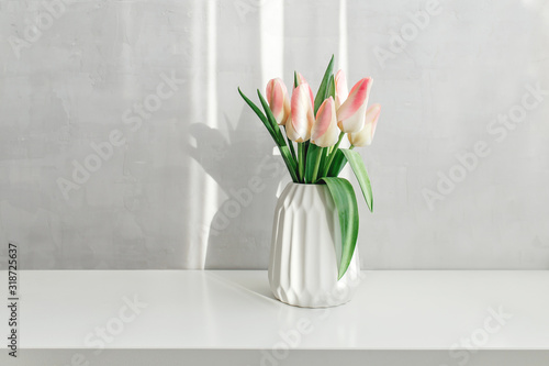 Fototapeta Naklejka Na Ścianę i Meble -  Light pink tulips in a white geometric ceramic vase stand on a white table near grey wall. Bouquet of flowers in the morning sun beams.