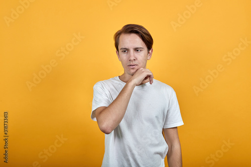 A handsome young man is thinking propping his chin with his hand in white t shirt on yellow background