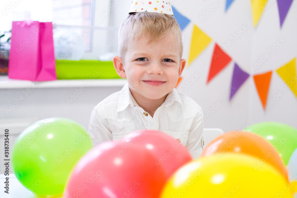 Portrait of blonde caucasian boy smiling at camera near birthday rainbow cake. Festive colorful background with balloons. Portrait of blonde caucasian boy smiling at camera near birthday rainbow cake. Festive colorful background with balloons.