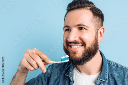 A young handsome man with a beard is brushing his teeth on an isolated blue background with raised