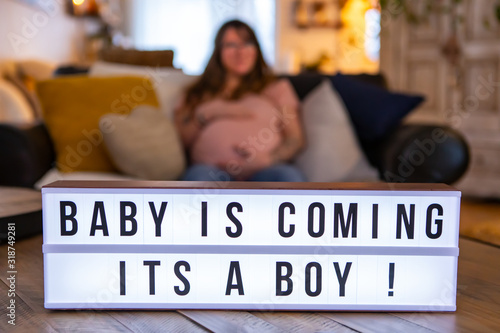 A selective focus shot on a cinematic LED lightbox with the message baby is coming its a boy, a blurry pregnant woman is seen sitting in background