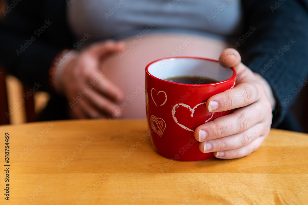 A selective focus view of a pregnant girl sitting at a wood table with cup of coffee in novelty valentine mug, a blurry hand is seen feeling baby kick A selective focus view of a pregnant girl sitting at a wood table with cup of coffee in novelty valentine mug, a blurry hand is seen feeling baby kick