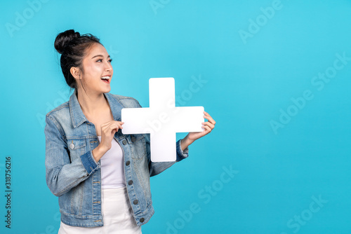 Happy asian woman standing and holding plus or add sign on blue background. Cute asia girl smiling wearing casual jeans shirt and showing join sign for increse, upgrade and more benefit concept photo