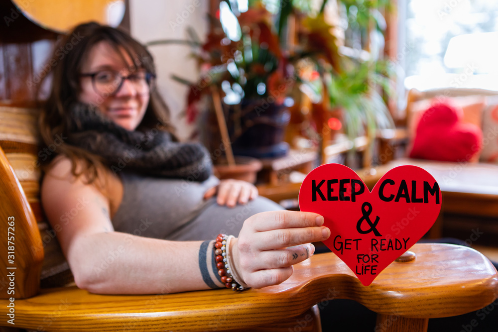A selective focus view of a heavily pregnant woman holding a red heart sign saying keep calm and get ready for life, close to labor at valentine A selective focus view of a heavily pregnant woman holding a red heart sign saying keep calm and get ready for life, close to labor at valentine
