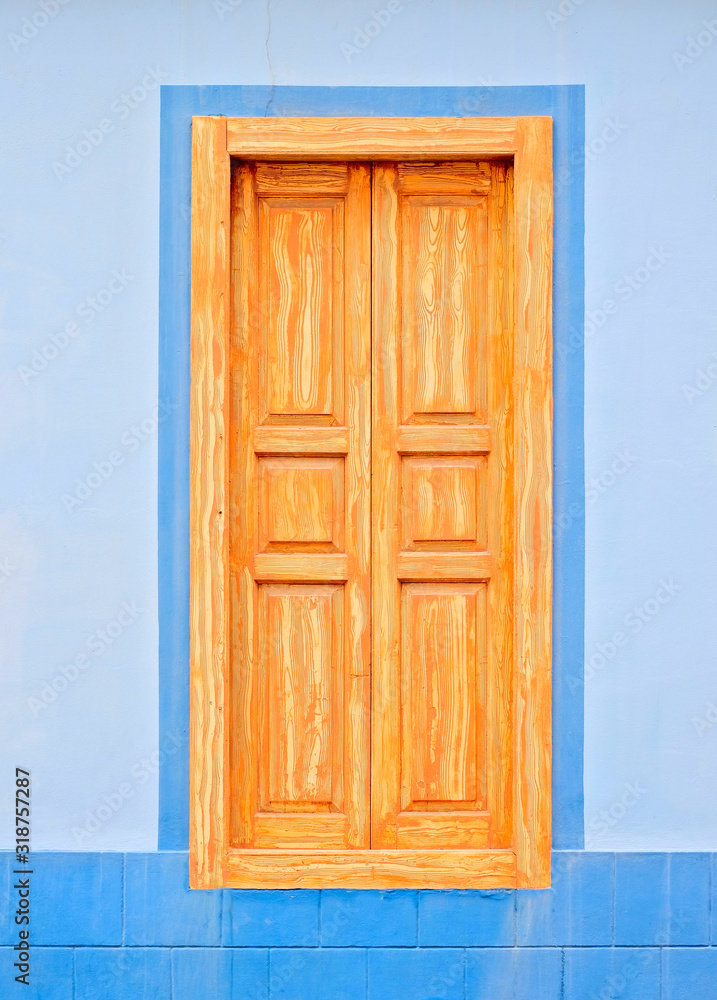 A tall window, closed, shutters in light timber color, light blue wall.