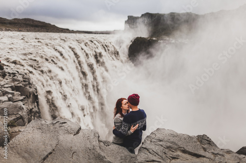 Young stylish couple in love kissing together near famous Icelandic landscape Dettifoss waterfall. Traditional wool sweaters, hat, red hair, gray skirt. Dramatic landscape, cold weather in Iceland.