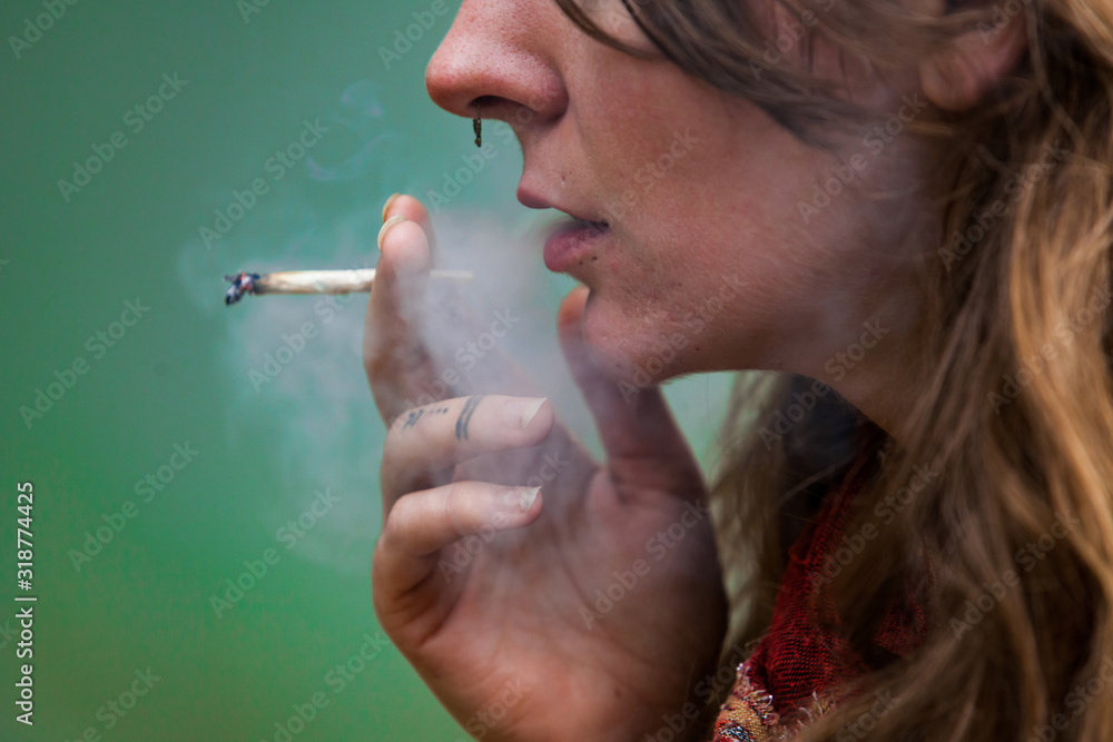 Obraz premium Closeup cropped shot of lower half of caucasian woman's face smoking hand rolled marijuana joint. Cannabis smoke being exhaled by pierced and tattooed woman.