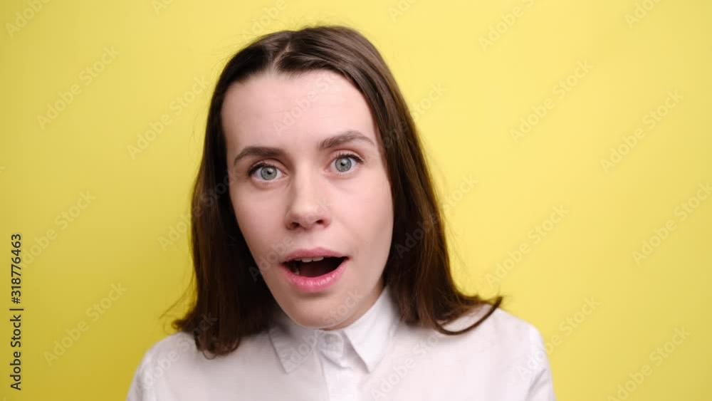 Stupefied shocked brunette woman keeps mouth widely opened, keeps hands on cheeks, jaw dropped and looks shock at camera, dressed in white shirt, standing isolated over yellow background. Stupefied shocked brunette woman keeps mouth widely opened, keeps hands on cheeks, jaw dropped and looks shock at camera, dressed in white shirt, standing isolated over yellow background.