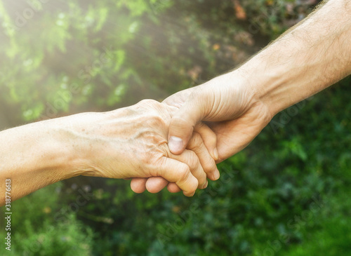 Senior and young hands outside closeup. Elderly woman and young man holding hands together, green blurred background, sunlight. Love, warmth, take care concept, Valentines, mothers day, donate, help.