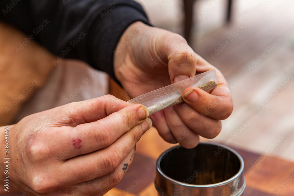 Selective focus closeup shot of tattooed hands twisting a paper with ground marijuana preparing a cannabis below a metal cylinder over a checkered table. Selective focus closeup shot of tattooed hands twisting a paper with ground marijuana preparing a cannabis below a metal cylinder over a checkered table.