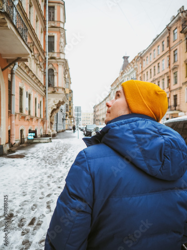 A man in a winter jacket and a bright yellow hat walks through the winter snow-covered St. Petersburg, along the beautiful historical streets