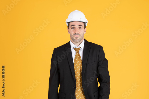 Young handsome businessman wears suit and construction helmet on isolated yellow background. © Karlie Studio