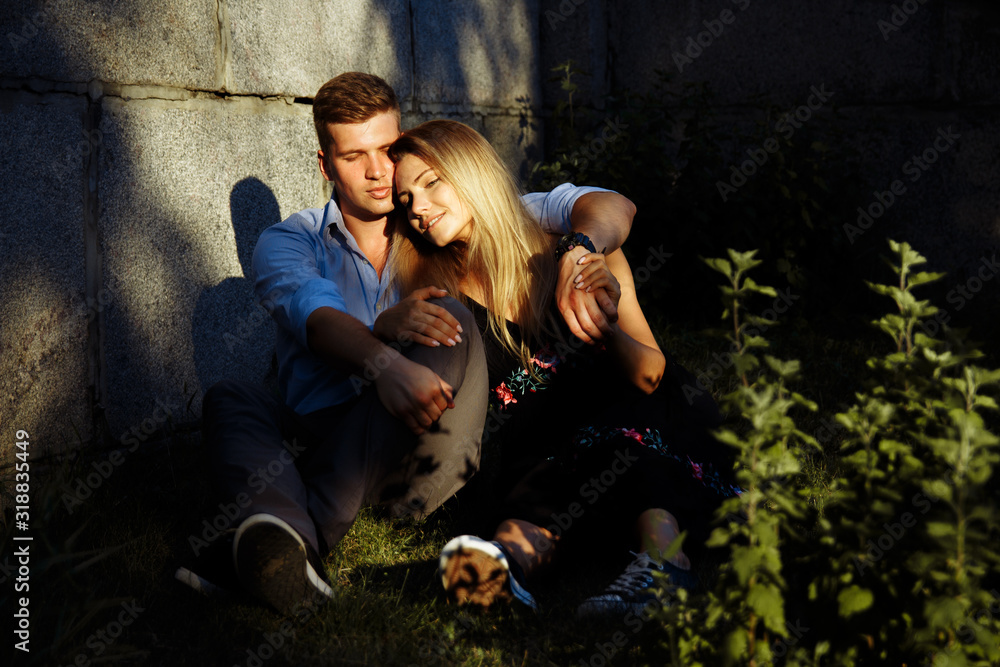 a couple in love is sitting on the grass near the wall, the sun is shining in the eyes, the guy is squinting, we are hugging a girl in a black dress