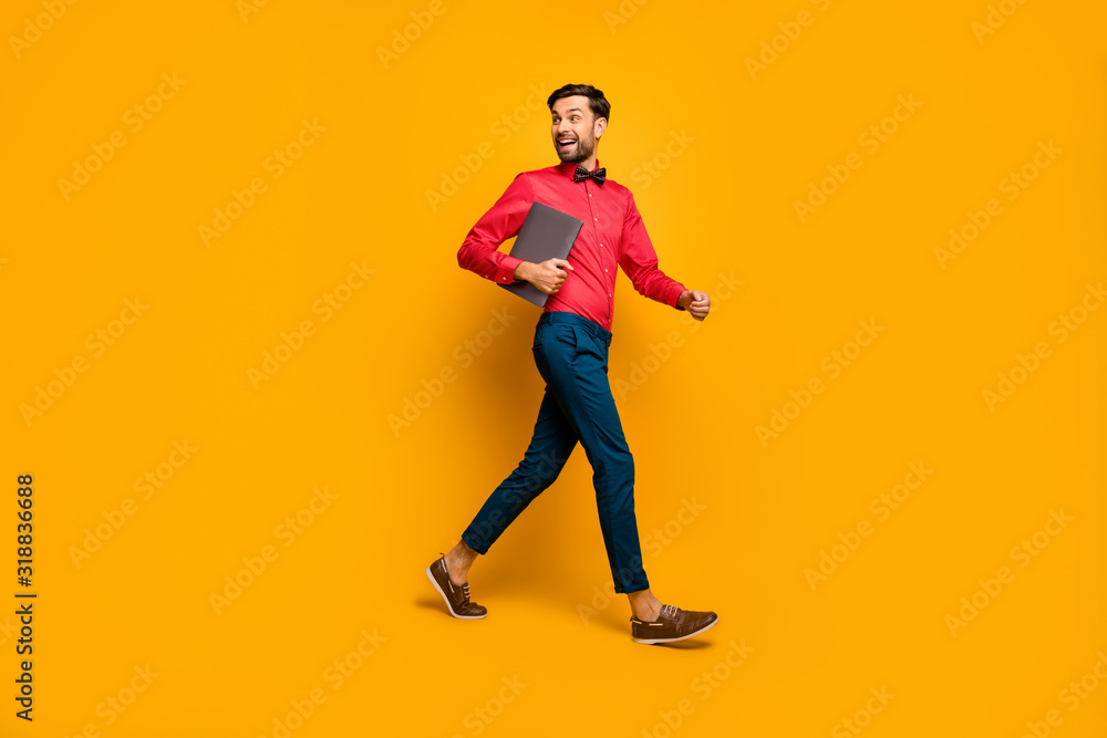 Full length profile photo of amazing business man walking corporate meeting training carry laptop wear stylish red shirt bow tie blue pants shoes isolated yellow color background Full length profile photo of amazing business man walking corporate meeting training carry laptop wear stylish red shirt bow tie blue pants shoes isolated yellow color background