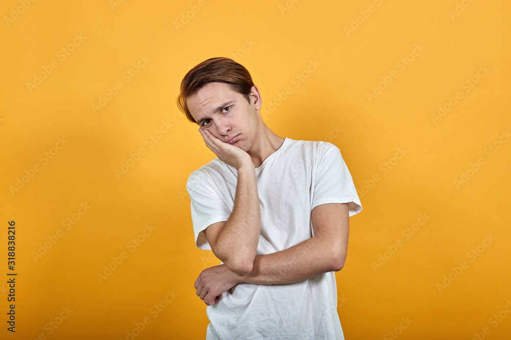 A young handsome man in white t-shirt looks distressed at camera with head on hand on yellow background A young handsome man in white t-shirt looks distressed at camera with head on hand on yellow background