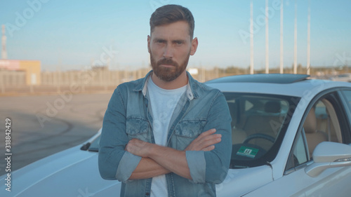 Portrait of severe confident guy leaning on car at parking space. Close-up handsome serious young man driver against modern auto under sunset.