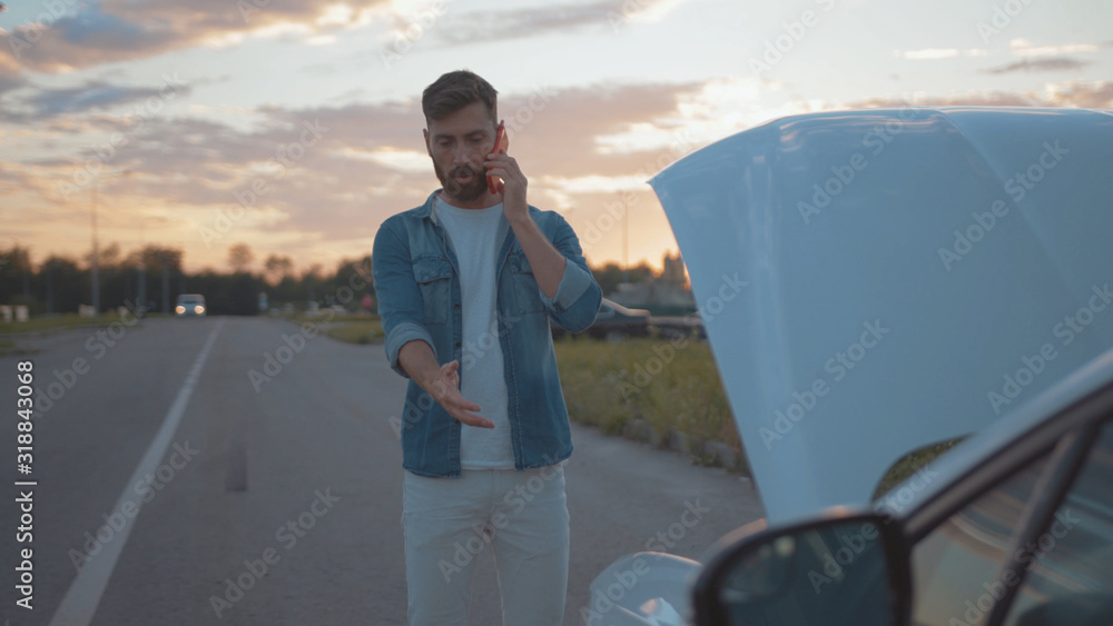 Nervous young man using smartphone calling car assistance services standing by breakdown automobile on the roadside at sunset.