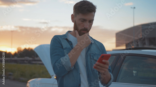 Nervous young man using smartphone calling car assistance services standing by breakdown automobile on the roadside at sunset.