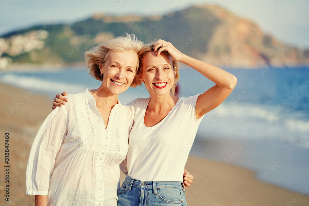 Outdoor portrait of smiling happy caucasian senior mother with her adult daughter hugging and looking at the camera on sea beach. Outdoor portrait of smiling happy caucasian senior mother with her adult daughter hugging and looking at the camera on sea beach.