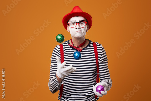 Fotografia Mime man in vest and red hat juggles with colorful balls on orange background