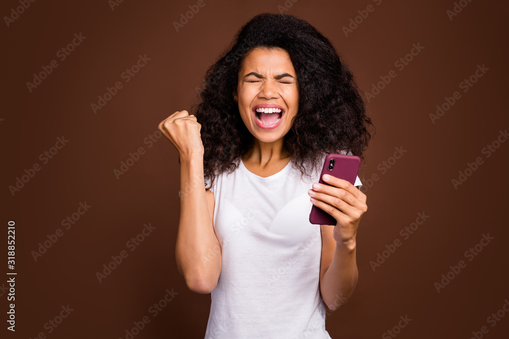 Portrait of delighted afro american girl use smart phone read social media news celebrate victory scram yes raise fists wear white t-shirt isolated over brown color background Portrait of delighted afro american girl use smart phone read social media news celebrate victory scram yes raise fists wear white t-shirt isolated over brown color background