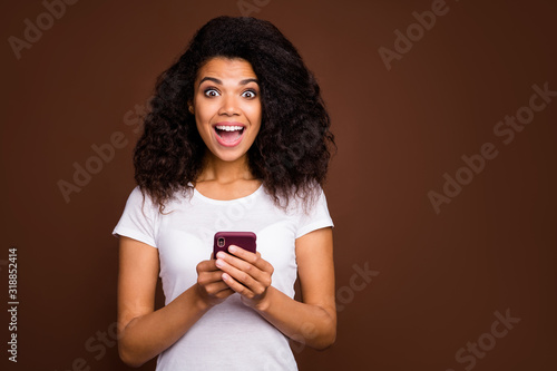 Portrait of amazed excited afro american girl use cell phone read social media news impressed scream wow omg wear trendy clothing isolated over brown color background