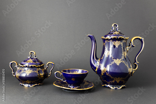 Closeup of a beautiful cobalt blue colored vintage porcelain tea set with golden floral pattern on dark gray background. The set includes a tea pot, a sugar bowl and a tea cup.