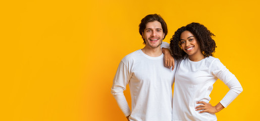 Smiling interracial couple cuddling while posing to camera over yellow background