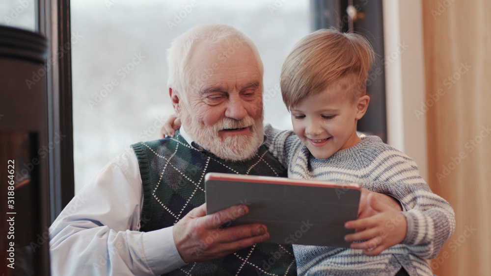 Smiling grandfather and grandson using digital tablet for surfing internet  and playing game near the fireplace at home grandpa adult grandchild child  childhood communication computer Stock-Foto | Adobe Stock