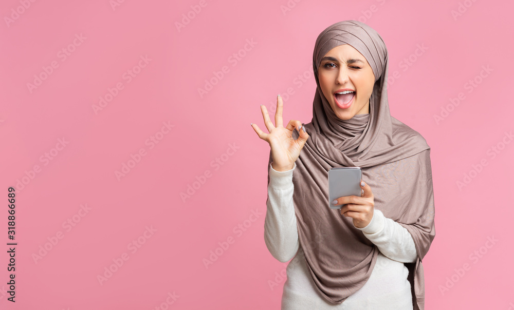 Arabic woman in hijab using smartphone, winking and showing ok gesture Arabic woman in hijab using smartphone, winking and showing ok gesture