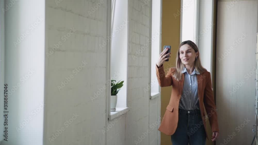 Smiling american businesswoman having video call and walking in office room. Happy female manager holding smartphone in hand, watching in camera with smile and talking. Elegant lagy going on Smiling american businesswoman having video call and walking in office room. Happy female manager holding smartphone in hand, watching in camera with smile and talking. Elegant lagy going on