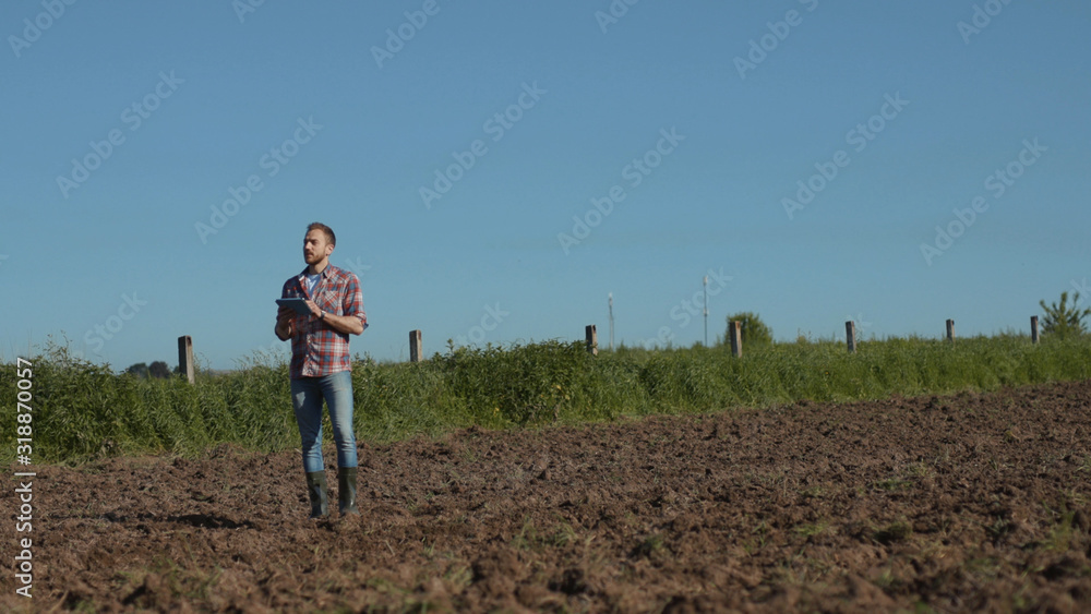 Beautiful young man agronomist working in the soil ground field using a tablet. Prosperous farmer checking soil and planted plants at a farmland in sunny weather in spring.