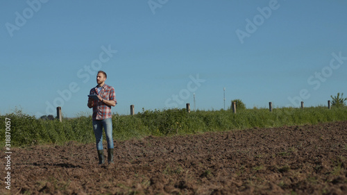 Beautiful young man agronomist working in the soil ground field using a tablet. Prosperous farmer checking soil and planted plants at a farmland in sunny weather in spring. © america_stock