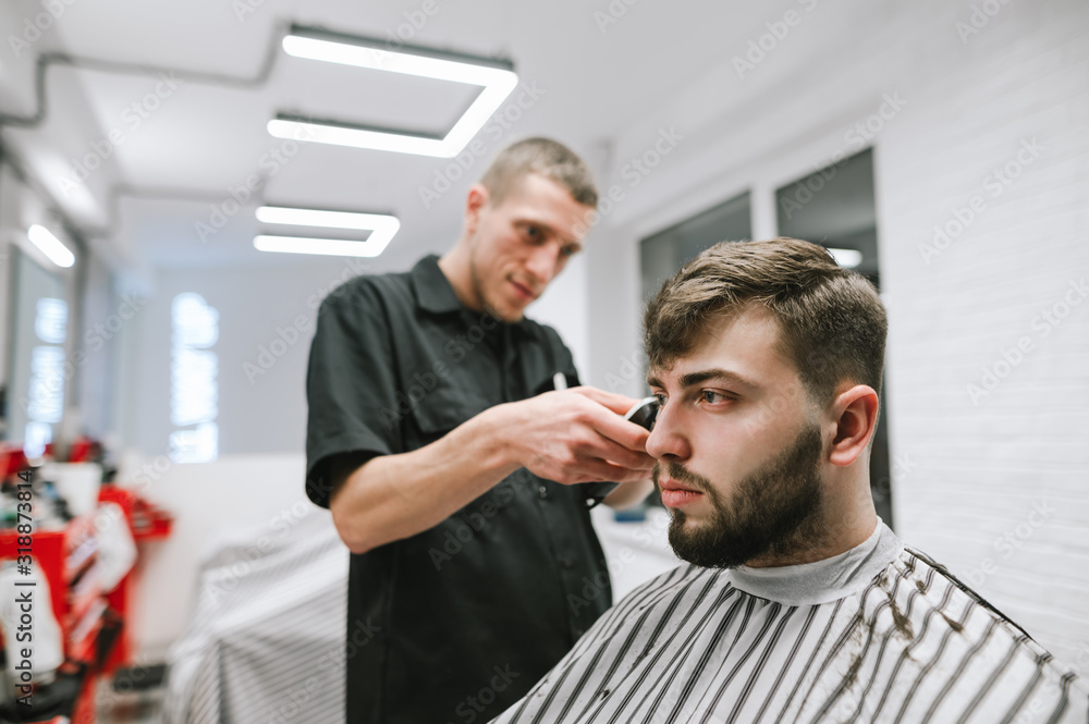 Obraz premium Portrait of a bearded male client sitting in a chair and crouching in a barber, looking away with a serious face. Man with beard and hair care in barbershop. Haircut in a professional barber.