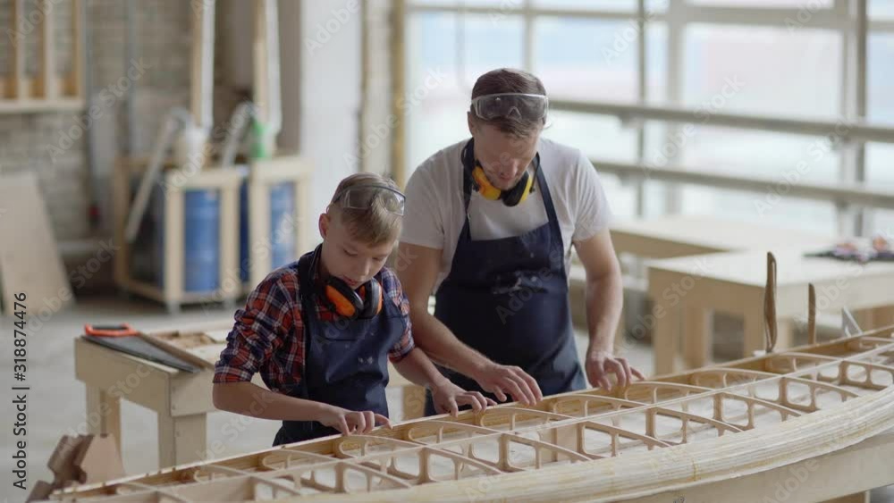 Carpenter and his little son standing together at workbench in woodworking shop. Man explaining kid how to build small wooden boat Carpenter and his little son standing together at workbench in woodworking shop. Man explaining kid how to build small wooden boat