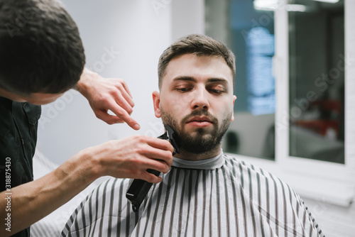 A professional male hairdresser does a beard haircut in a handsome man, uses a clipper. Attractive makes a beard and head hairstyle in a barber shop, close-up portrait.
