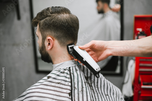 Professional male hairdresser does hairstyle to bearded man in barber shop, back view, barber's hand with clipper. Barber clipping client in men's hairdresser, closeup photo