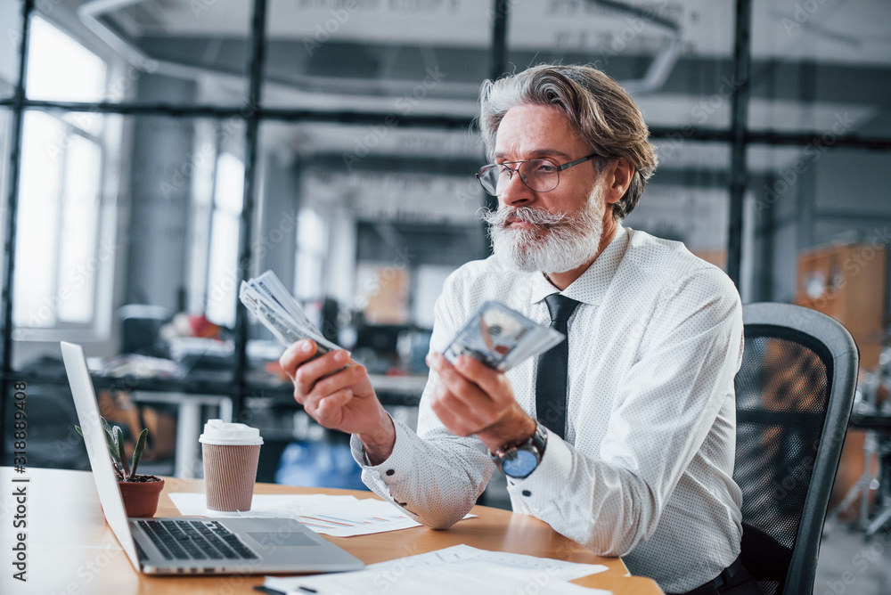 Happy mature businessman with grey hair and beard in formal clothes is in the office holding money Happy mature businessman with grey hair and beard in formal clothes is in the office holding money