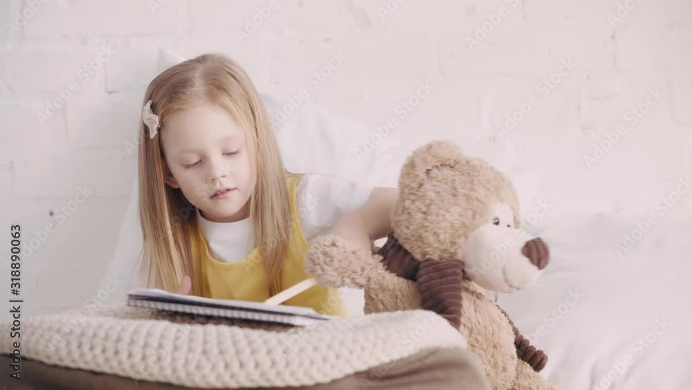 Child holding teddy bear while drawing on bed Child holding teddy bear while drawing on bed