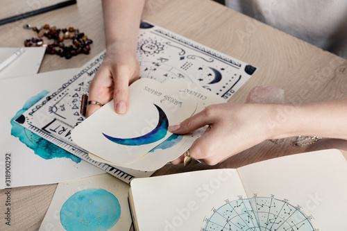 Cropped view of astrologer holding cards with watercolor drawings by birth chart with zodiac sign on table photo