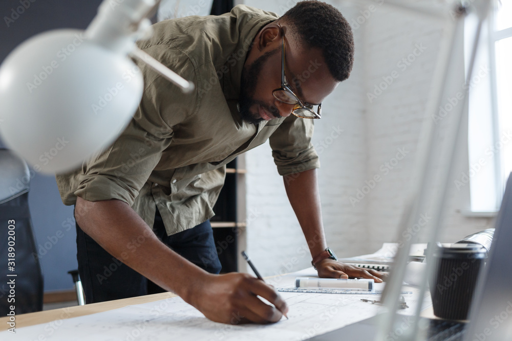 Afro-American architect working in office with blueprints.Engineer inspect architectural plan, sketching a construction project. Portrait of black handsome man sitting at workplace. Business concept. Afro-American architect working in office with blueprints.Engineer inspect architectural plan, sketching a construction project. Portrait of black handsome man sitting at workplace. Business concept.