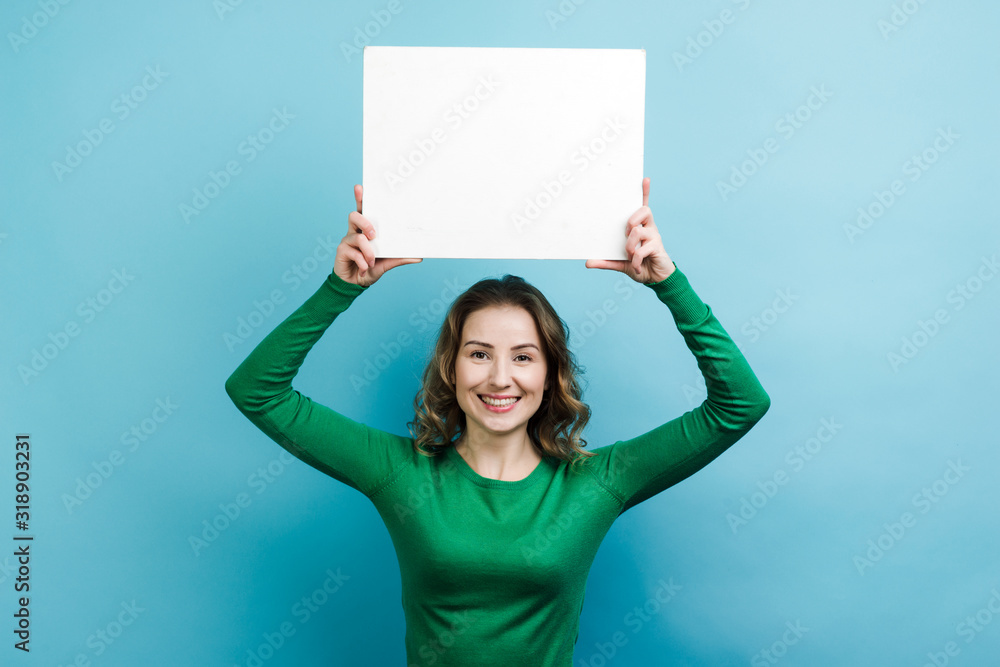 Curly girl wearing in green sweater holding a white board copy space above her head against blue background Curly girl wearing in green sweater holding a white board copy space above her head against blue background
