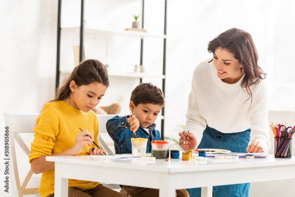 happy woman standing near table and drawing with paints together with children happy woman standing near table and drawing with paints together with children