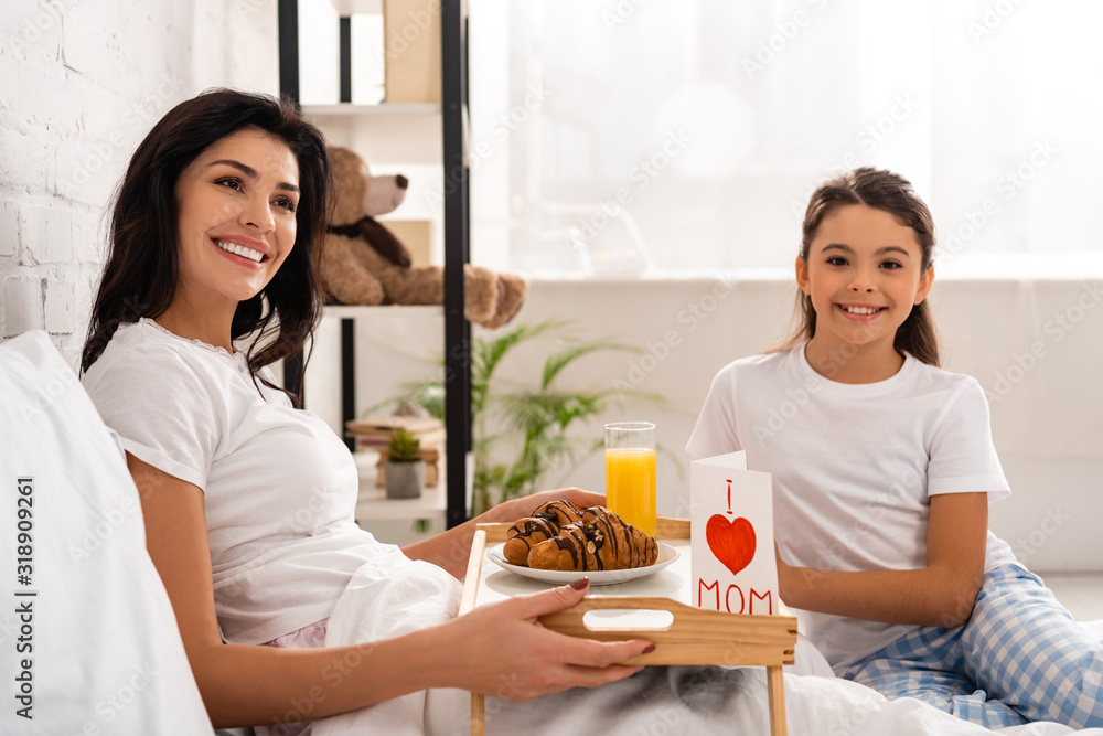 adorable child sitting near mother holding tray with breakfast, mothers day card with heart sign and mom lettering adorable child sitting near mother holding tray with breakfast, mothers day card with heart sign and mom lettering